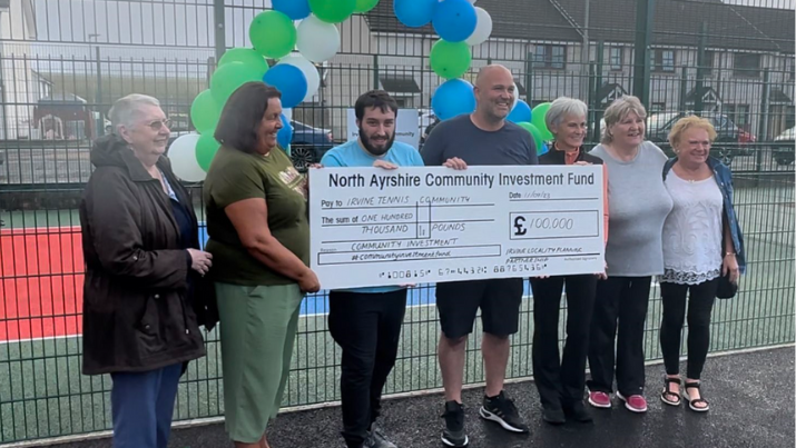 Group of people holding photo prop cheque outside of revamped tennis court with famous tennis coach Judy Murray