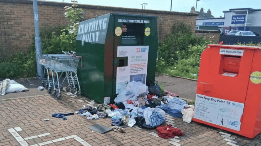 Two charity clothing banks, with shops in the background and burst bags of donations strewn in the foreground